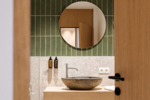 Apartment-Badezimmer im Hideaway Oetz – Waschtisch aus Eiche mit Waschbecken aus Naturstein, grüne Stäbchenfliesen und runder Spiegel
