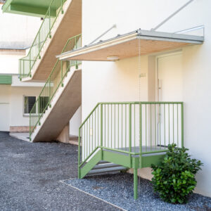 Lebe'scharnitz – Außentreppe und Wohnungszugang Lebe'scharnitz: Wohnungseingang mit Vordach und grünem Stahlgeländer, dahinter die neue Außentreppe aus Sichtbeton