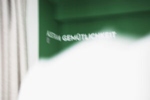 Signage – Austrian Gemütlichkeit Weißer Schriftzug „Austrian Gemütlichkeit" auf einer dunkelgrünen Wandnische, davor ein unscharfer heller Vordergrund