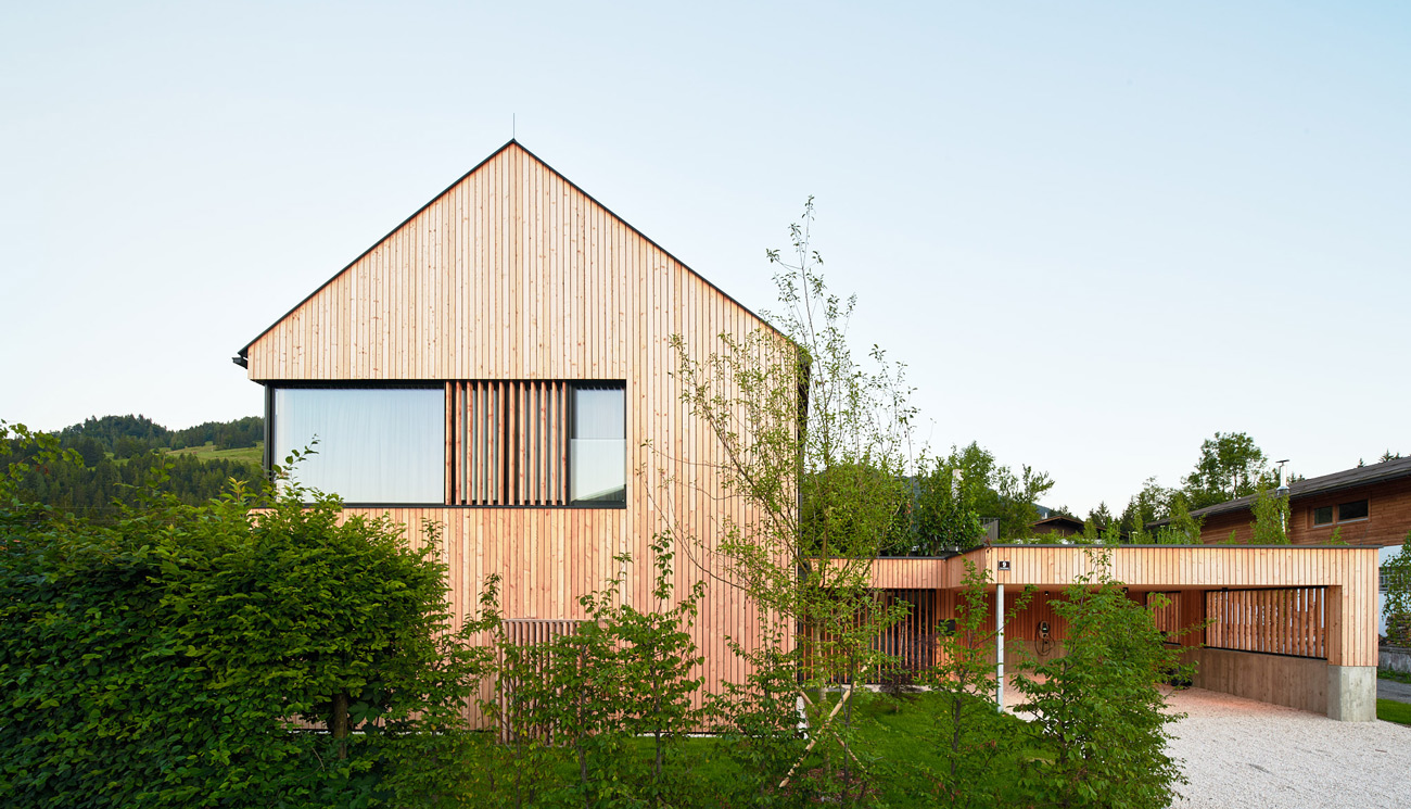 Moderner Holzbau mit Satteldach und senkrechter Lärchenholzfassade, großem Fenster und angebautem Carport, umgeben von Bäumen und Hecken im Grünen